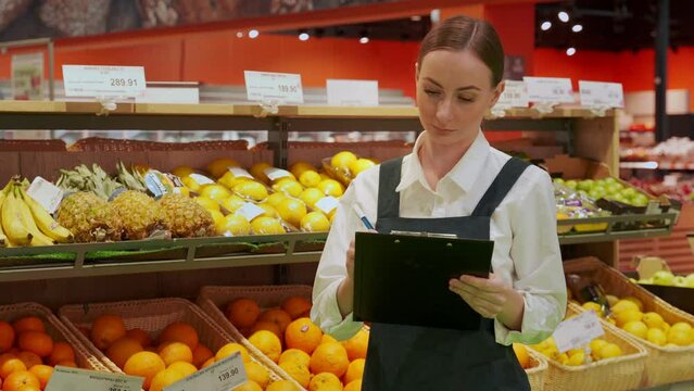 Young store manager examines condition of fruit products and writes note on notebook. Female worker checks implementation date in greengrocer department closeup