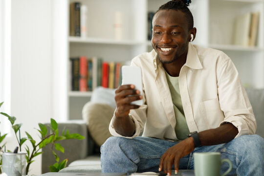  African American Man In Headphones Using A Smartphone, Browsing The Internet While Sitting On The Sofa At Home, Writing On A Mobile Phone Or Using A New Application Indoors