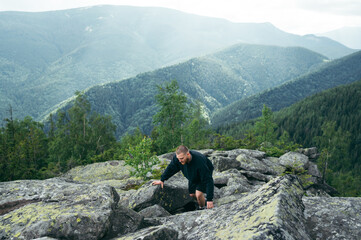 A male tourist climbs a rock on top of a mountain against the background of beautiful views of the Carpathians. Active recreation in the mountains.