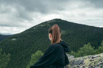 Back view of female hiker in casual clothes sitting on rock on top of mountain and looking at mountain landscape.