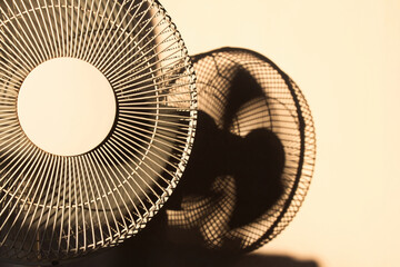 An electric fan with blurred blades moving casting a blurry shadow on the plain white wall in the...