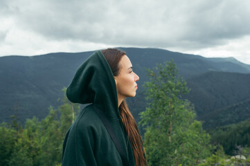 Portrait of a cute female tourist in a hoodie in the mountains against the background of beautiful views, looking away with a serious face. Woman on a hike.