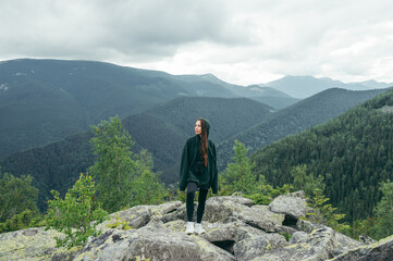 A woman tourist in casual clothes stands on a stone on top of a mountain against the background of beautiful views and looks away. Active recreation.