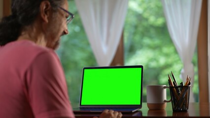 Over shoulder view of mature man making video call on laptop green screen. Senior man talking and...