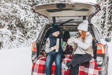 Two friends teenagers boy and girl with thermos sitting in trunk of car decorated for Christmas. Road trip adventure and local travel concept. Winter picnic in snowy forest. New year vacation.