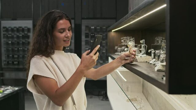 Waist-up of young brown-haired woman taking picture with smartphone of earrings in contemporary jewelry store