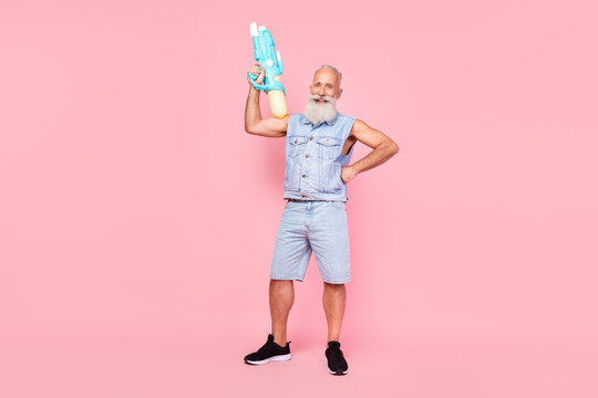 Full length photo of attractive strong man pensioner dressed denim vest rising water gun isolated pink color background