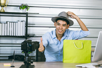 A young Asian influence male vlogger thumbs up while holding a shopping bag while live streaming,...