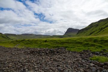 Wide angle views of Talisker Bay, Isle of Skye, Scotland, with its rocky beach, black stones, green fields, and a waterfall at the end of beach. Scattered clouds on blue sky, summer scottish weather.