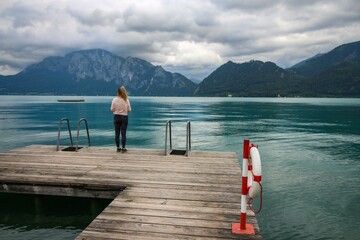 Attersee - famous Austrian turquoise lake. The view on the woman standing on the wooden pier looking at the lake and beautiful cloudy mountains in the background. 