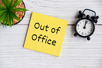 Out of office text on sticky note with alarm clock and plant background. Business concept.
