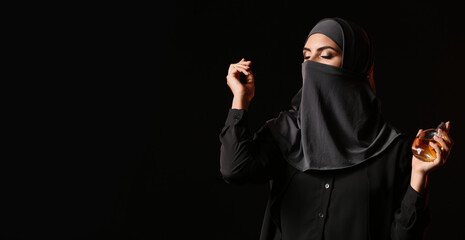 Beautiful Muslim woman holding bottle of perfume on dark background with space for text