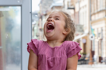 An emotional portrait of a small blonde 4-year-old girl catching soap bubbles on a blurry background of the old city in summer.