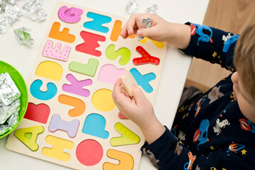 Two year boy playing with wooden alphabet letters board. Letters wrapped in foil. Intellectual game, preschool implement for early education. Verbal and memory training exercise.