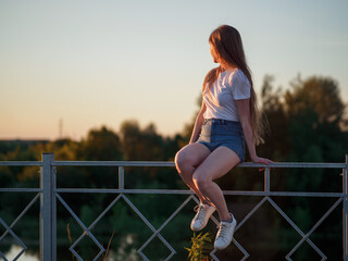 A pretty young woman in a white blouse and shorts is sitting on the railing, enjoying the summer sunset and freedom.