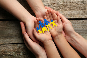 Fototapeta premium Parents and child holding paper family cutout in colors of Ukrainian flag at wooden table, top view