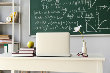 Apple, books, laptop, eyeglasses and vase with flower on table in classroom
