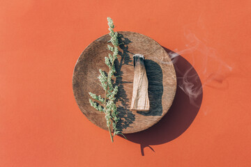 Burning palo santo stick with smoke on wooden stand over red orange background, burning incense....