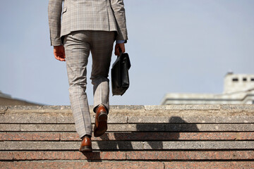 Man in a business suit with briefcase climbing stone stairs, male legs in motion on the steps. Concept of career, success, moving to the top, official or businessman - obrazy, fototapety, plakaty