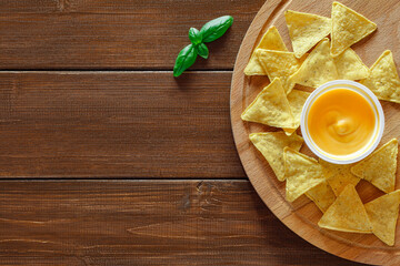 Nachos on round wooden stand, cheese salsa and green basil leaf on brown plank dark background, top view, space to copy text..