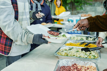 Close-up of volunteers serving homeless people and putting meal on plates, they feeding poor people...