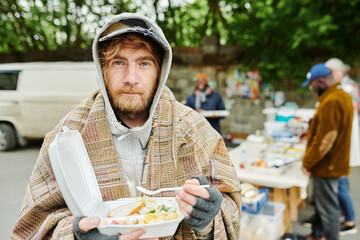 Portrait of bearded homeless man in warm clothing looking at camera while eating food outdoors...