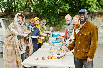 Portrait of group of volunteers and homeless people smiling at camera while arranging charity for...