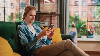 Obraz premium Young Handsome Adult Man Sitting on a Couch in Living Room, Relaxing and Using Smartphone. Creative Male Checking Social Media, Chatting with Friends, Browsing Internet. City View from Big Window.