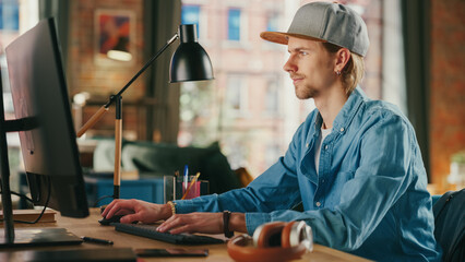 Young Handsome Man Working from Loft Apartment on Desktop Computer. Creative Male Checking Social Media, Browsing Internet from Home. Urban City View from Big Window.