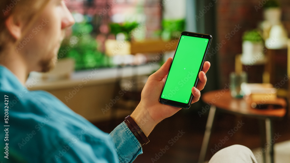 Leinwandbilder man holding a smartphone with green screen mock up display. male is resting at home, watching videos