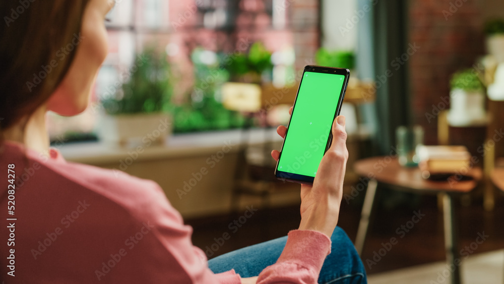 Poster feminine hand holding a smartphone with green screen mock up display. female is relaxing at home, wa