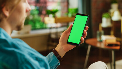 Man Holding a Smartphone with Green Screen Mock Up Display. Male is Resting at Home, Watching...