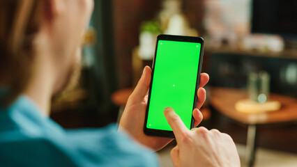 Man Scrolling Feed on Smartphone with Green Screen Mock Up Display. Male Having Fun and Enjoying Content on Social Media on Mobile Device while Resting at Home. Close Up Over the Shoulder Photo.