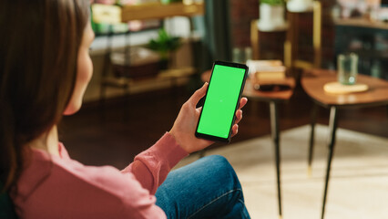 Feminine Hand Holding a Smartphone with Green Screen Mock Up Display. Female is Relaxing at Home, Watching Videos and Reading Social Media Posts on Mobile Device. Close Up Over the Shoulder Photo.