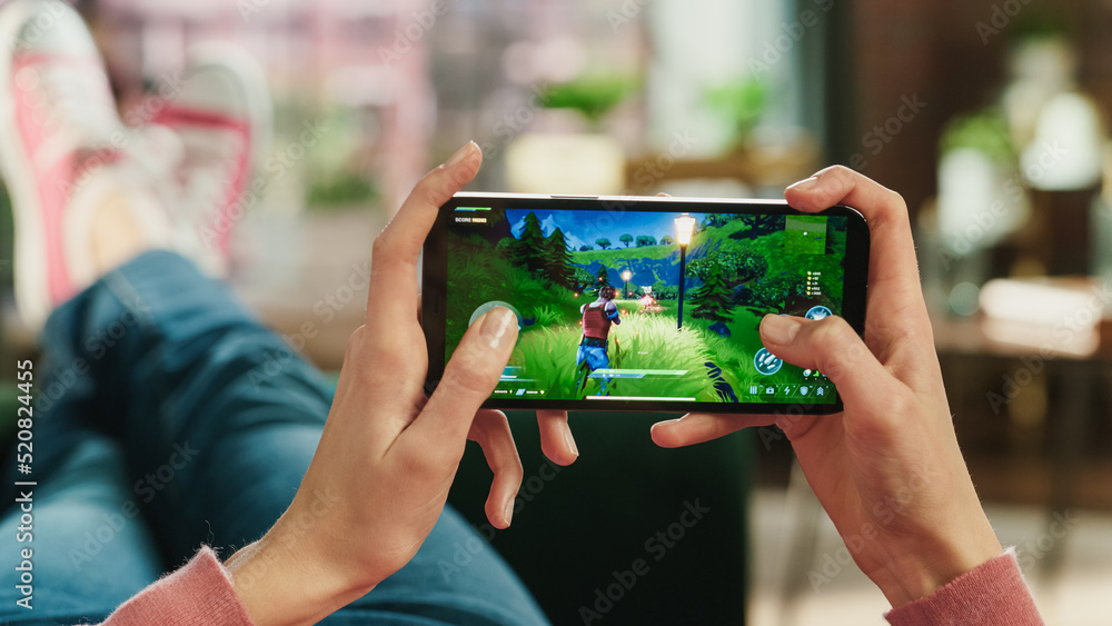 Poster female is relaxing on a couch at home, playing an interactive pvp shooter video game on her smartpho