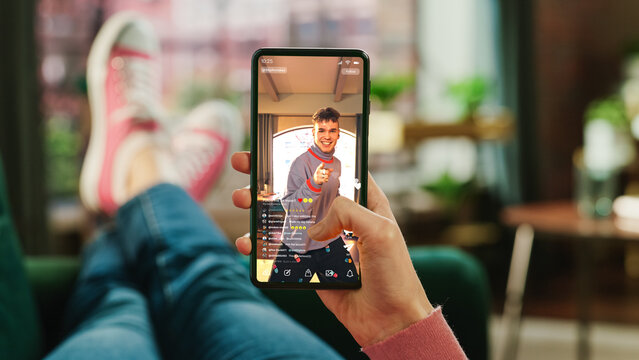 Feminine Hand Scrolling Social Media Feed on Smartphone Display. Female is Relaxing on Sofa at Home, Watching Videos and Reading Social Network Posts on Mobile Device. Close Up POV Photo.