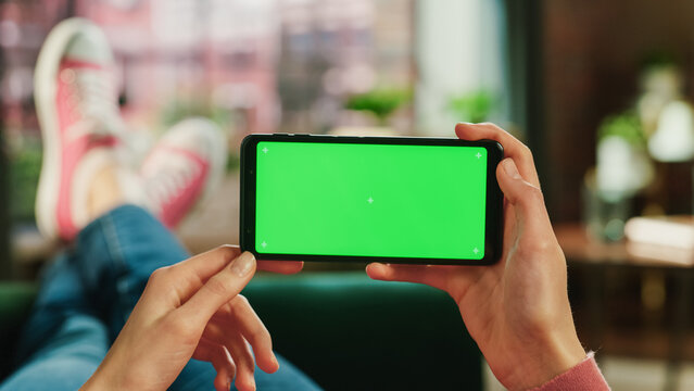 Woman Horizontally Holding a Smartphone with Green Screen Mock Up Display. Female is Relaxing on a Couch at Home, Watching Videos and Reading Social Media Posts on Mobile Device. Close Up POV Photo.