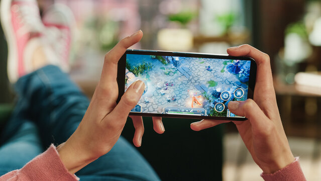 Female is Relaxing on a Couch at Home, Playing an Interactive PvP RPG Strategy Video Game on Her Smartphone. Gamer Lies on a Sofa in Living Room. Close Up POV Photo of Mobile Device Screen.