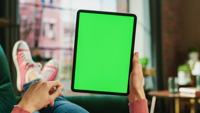 Feminine Hand Scrolling Feed on Tablet with Green Screen Mock Up Display. Female is Relaxing on Sofa at Home, Watching Videos and Reading Social Media Posts on Mobile Device. Close Up POV Photo.