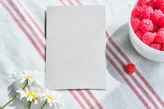 Greeting mockup scene on striped cotton napkin. Blank paper, cup with raspberries and daisy flowers
