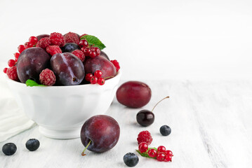 Plums, raspberries, red currants, blueberries in a white plate and on a light table.