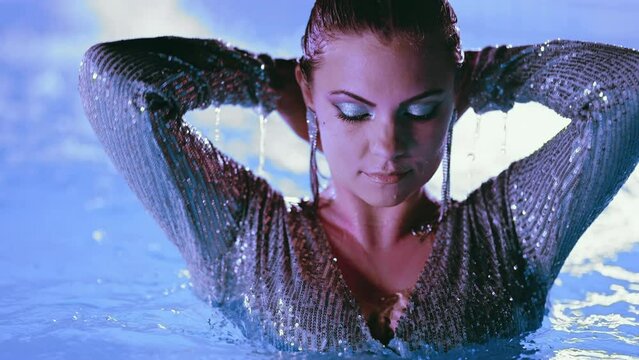 Beautiful woman posing in pool water under neon color light. Party, attractive chic in shining dress enjoying night time. Rich lifestyle, luxury life, influential people