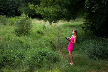 Naklejka na ściany i meble A slender young smiling freelancer girl with long blond hair, in a pink dress, stands in the forest on a green lawn and remotely works and studies on a tablet.