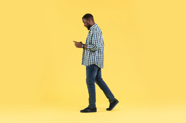 Black Male Using Smartphone Browsing Internet Walking Over Yellow Background