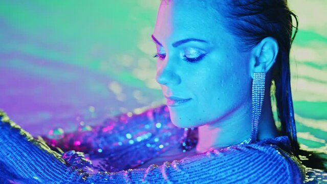 Beautiful woman posing in pool water under neon color light. Party, attractive chic in shining dress enjoying night time. Rich lifestyle, luxury life, influential people