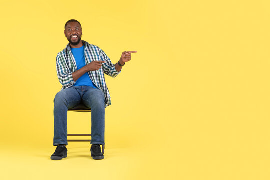 African American Male Pointing Fingers Aside Sitting On Yellow Background