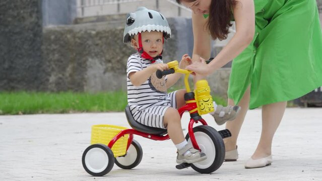 Mother teaching little child to ride tricycle for kids outdoors, toddler boy in funny bike helmet having fun playing in summer park. Outdoor playground riding activity for children.