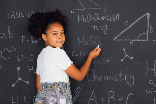 Happy African American schoolgirl solving problems near the blackboard at school, back to school concept.