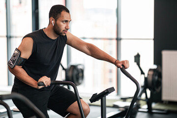 Sporty Young Black Man Using Elliptical Bike Machine While Training At Gym