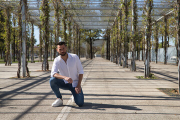Obraz premium Handsome young man with beard, wearing white shirt and jeans, with one knee on the ground and hands on legs. Concept fashion, beauty, modern, trend, model.